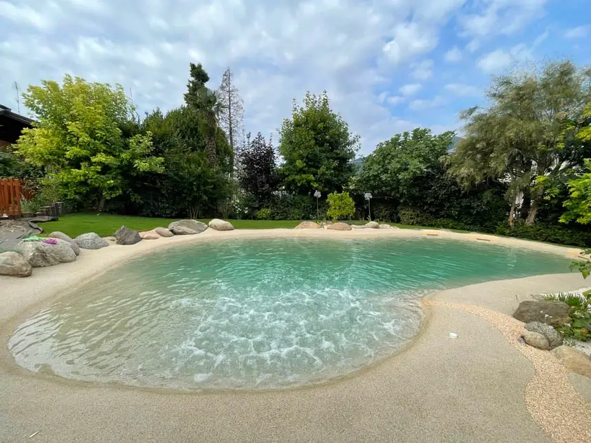 Beach-entry sand pool with clear turquoise water, framed by boulders and a lush garden